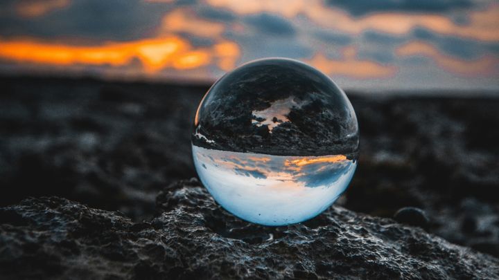 Photo of a marble sitting on dark rock, the top half reflecting the rock and the bottom half reflecting a light blue sky with clouds as the sun sets