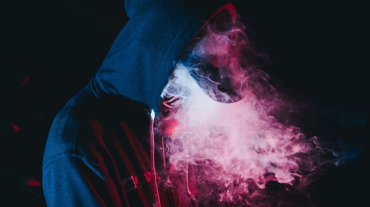 Photo of a hooded person exhaling smoke, lit to look pink, against a dark black background